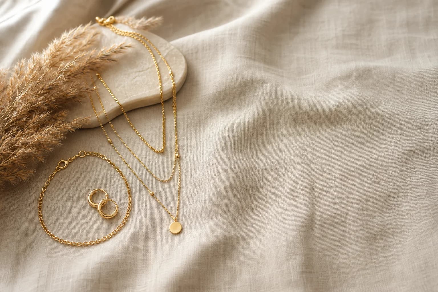 Layered gold necklace, chain bracelet and hoop earrings styled on warm linen